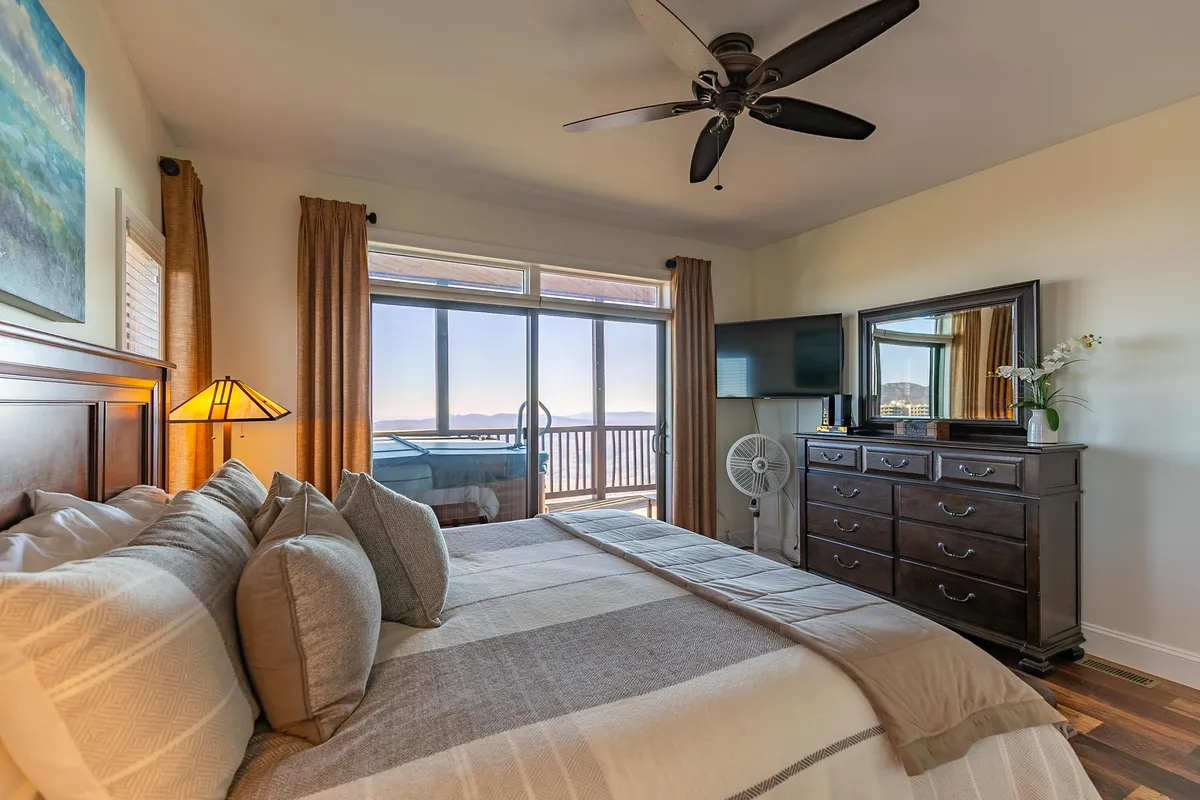 A tranquil bedroom featuring a bed with decorative pillows set against a large window that frames picturesque mountain views. The room includes a dresser with a TV and a ceiling fan, enhanced by soft, ambient lighting that creates a restful atmosphere.