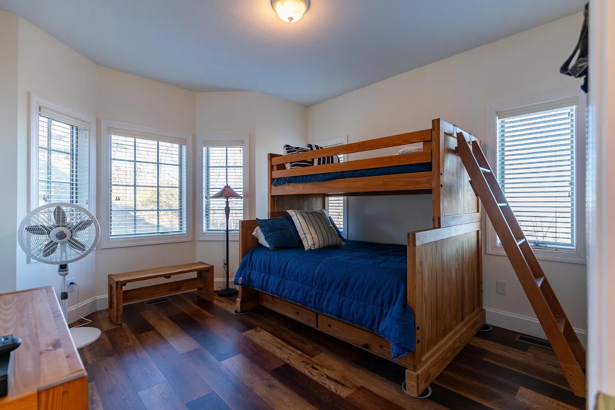 A cozy second bedroom with a twin-over-full bunk bed, wooden floors, a window with blinds, and a ceiling fan. The space feels inviting and family-friendly, perfect for both children and adults.