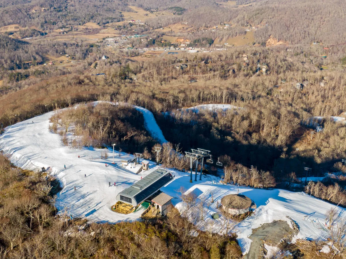 A sweeping exterior view featuring snow-covered ski slopes, a ski lift in action, the surrounding mountains, and a small lodge at the base. The landscape is bustling with winter activity, capturing the thrill and accessibility of ski-in/ski-out adventures.