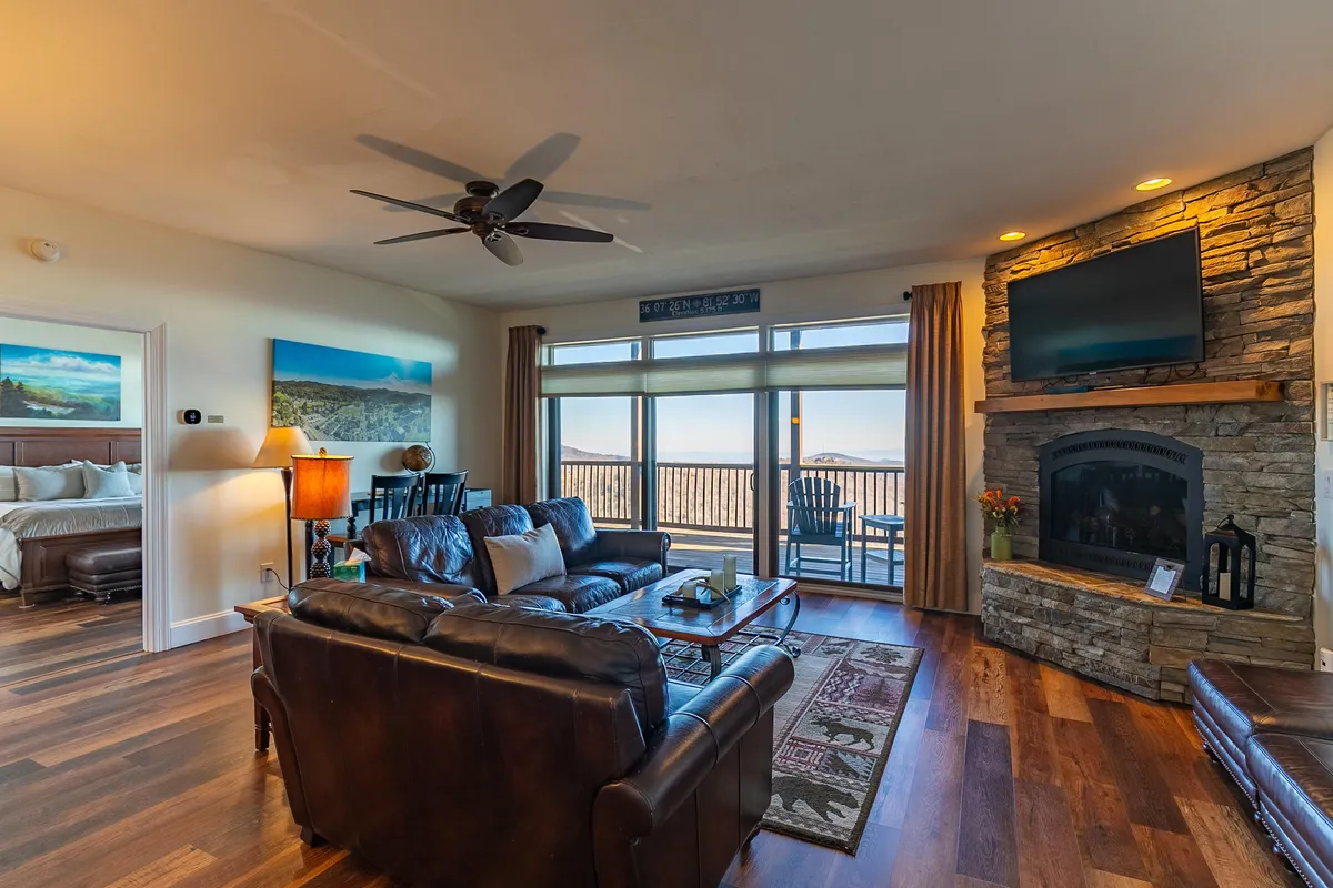 A warm, inviting living room featuring plush leather sofas arranged around a striking stone gas fireplace. A large TV sits above the mantle, while expansive windows offer a view of the outdoors and fill the space with natural light. A decorative rug and tasteful decor create a cozy yet stylish ambiance perfect for relaxing or gathering after an adventure.