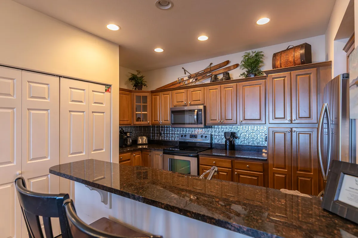 A well-equipped kitchen with a granite countertop, wooden cabinets, modern stainless steel appliances, and a row of kitchen bar stools. Decorative skis on the wall nod to the mountain setting, and the counter invites casual breakfasts or socializing while cooking.
