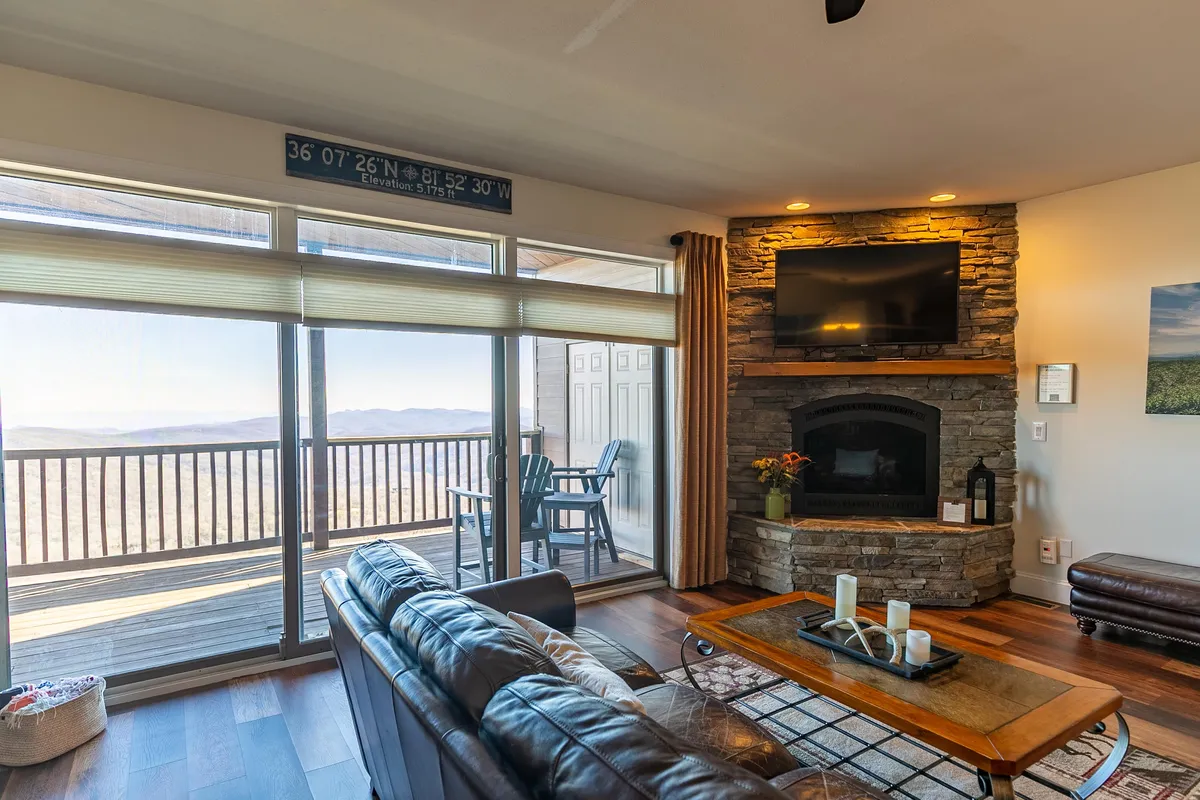 A cozy living room is shown with a large stone fireplace as its centerpiece, comfortable leather sofas, and expansive windows framing mountain vistas. The glow of the fire creates a warm and welcoming atmosphere perfect for relaxing after outdoor pursuits.