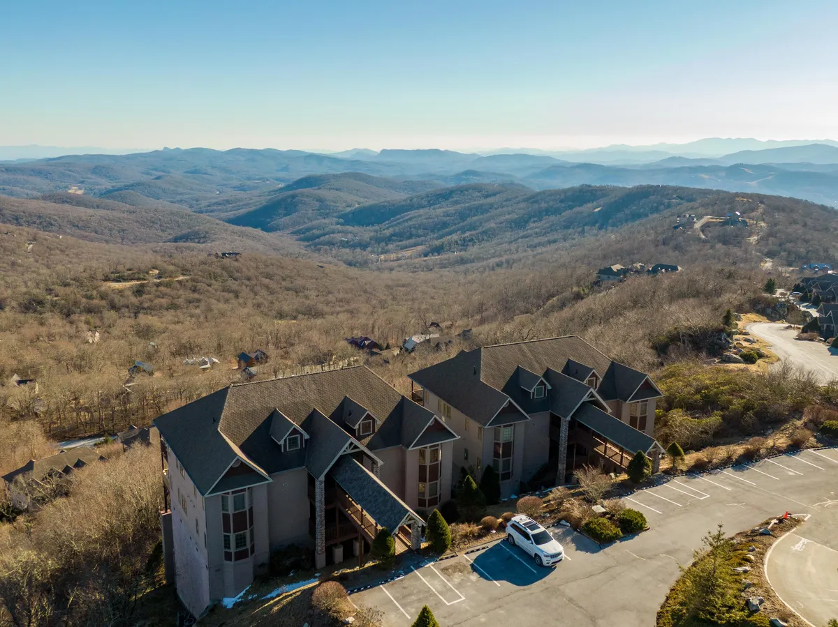 A tranquil exterior shot featuring the property with the majestic mountains in the background. The image includes the building structure nestled amidst nature, a parking area, and hints of the surrounding landscape. The natural lighting emphasizes the serene atmosphere, perfect for a romantic escape.