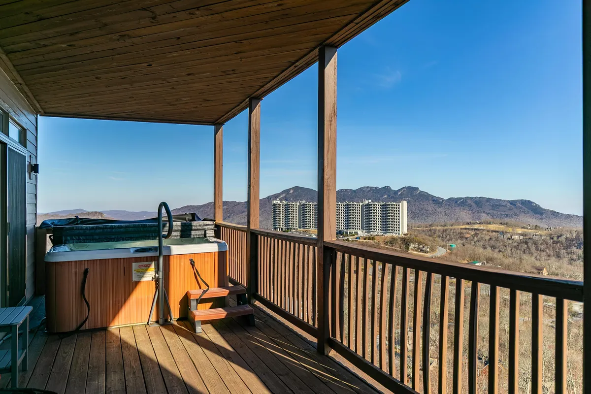 An inviting exterior space with a private hot tub overlooking majestic mountain views, framed by wooden railings. The scene suggests relaxation and après-ski comfort, with hints of sunset lighting.