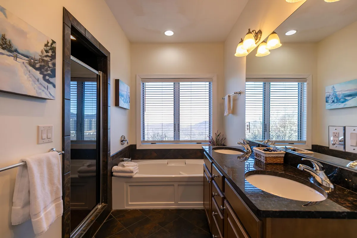 A spacious, serene bathroom with a modern bathtub positioned beneath large windows, double sinks with granite vanities, and tasteful artwork. The natural light enhances the inviting atmosphere, suggesting a space ideal for relaxation and self-care.