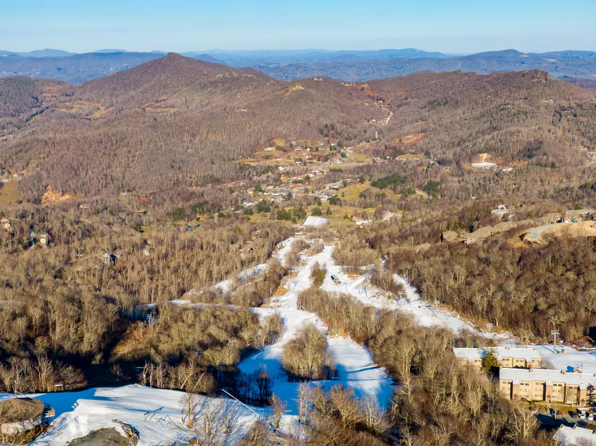 A panoramic winter landscape displaying mountain ranges, snow-draped forests, a small town in the valley, and a winding river. The scene is tranquil yet vibrant, capturing the essence of outdoor fun and relaxation.